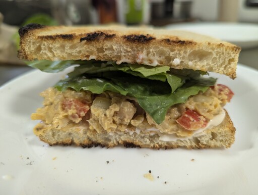 image contains a sandwich with toasted bread, lettuce, and a chickpea salad filling on a white plate and blurred background.